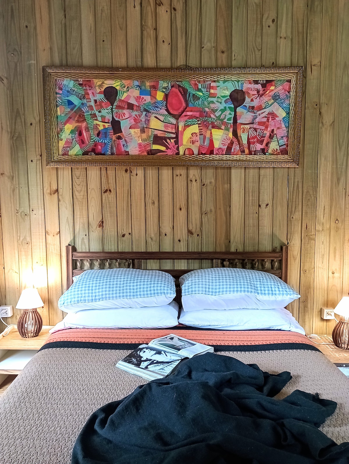 A cozy sleeping area features a wooden wall backdrop and a colorful artwork above the bed. Soft bedding is arranged on a spacious bed, with bedside tables complemented by lamp lighting. A black throw blanket and a magazine rest on the bed's edge, adding to the inviting ambiance.
