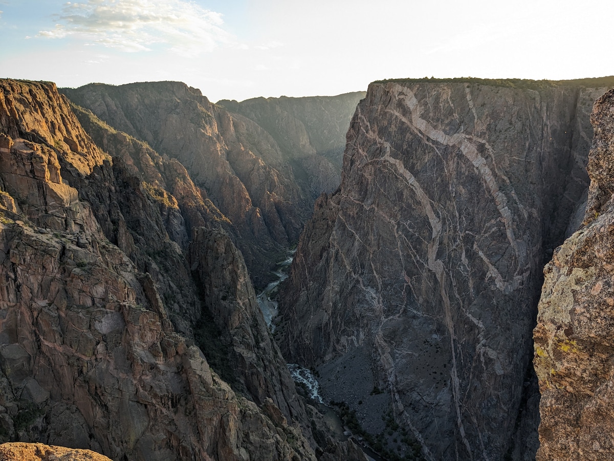 A breathtaking view of rugged canyon walls rises sharply from a narrow riverbed below. The interplay of light and shadow highlights the textured rock formations, with layers creating a striking contrast against the soft hues of the sky in the background.