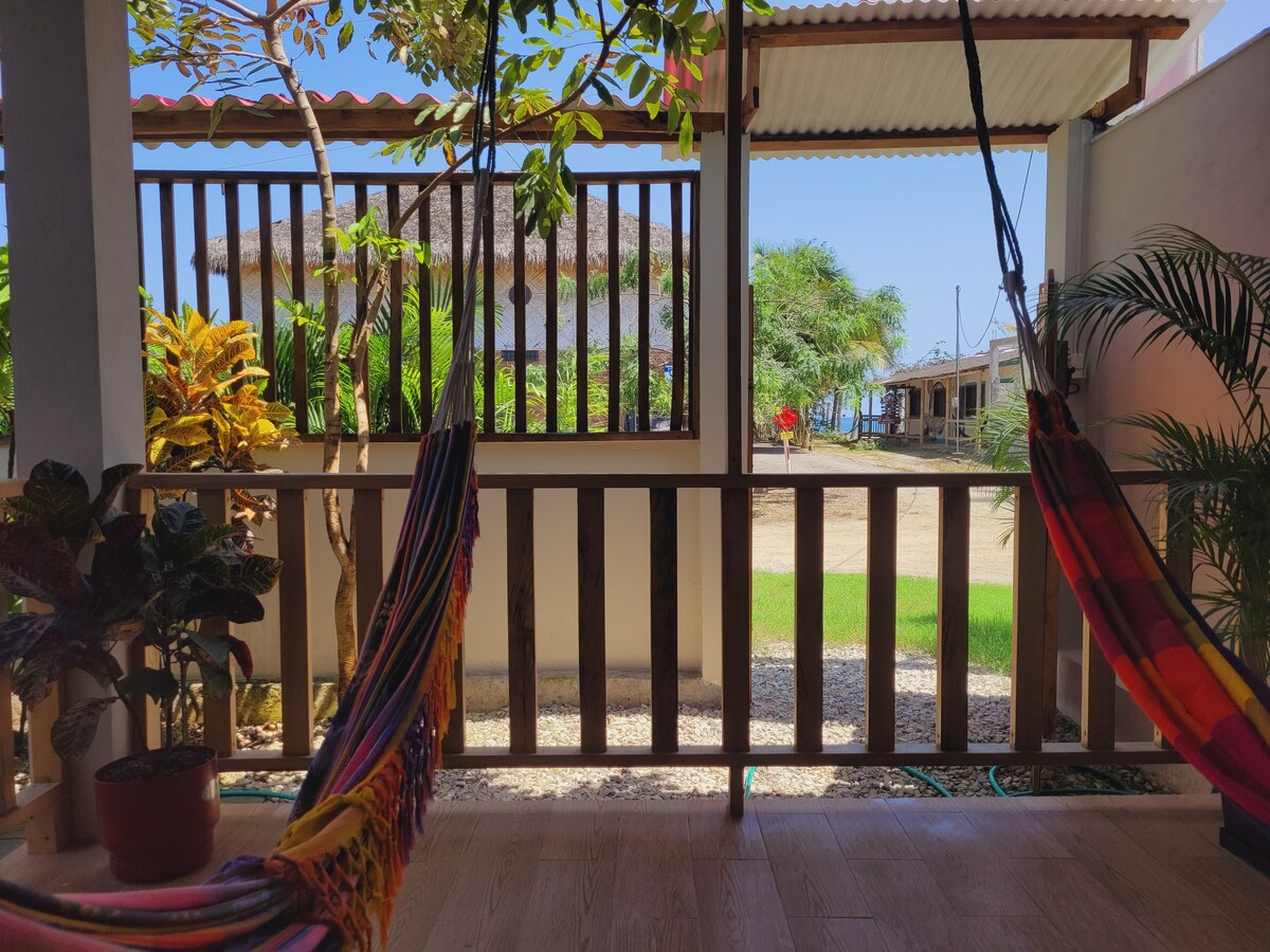 A shaded patio area is visible, featuring two colorful hammocks suspended between wooden posts. Lush green plants and trees are seen, providing a natural ambiance. The ocean can be seen in the background, with a hint of sandy shore and nearby structures.