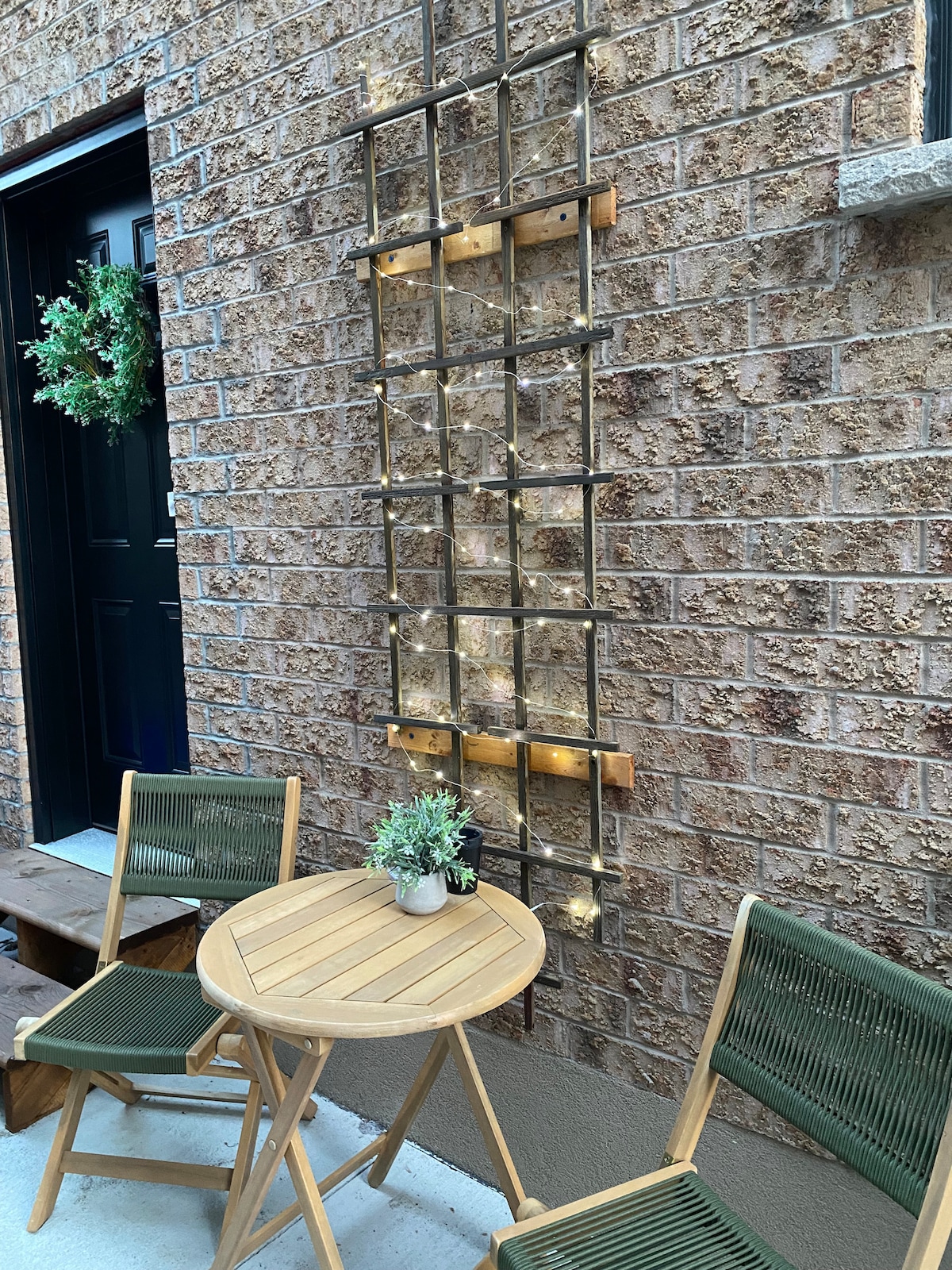 A small outdoor seating area features two green chairs positioned around a round wooden table. A decorative trellis with string lights is mounted on the brick wall, and a small potted plant rests on the table, creating a welcoming ambiance.