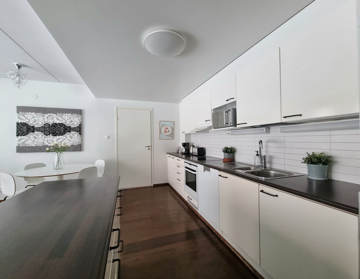 The kitchen features modern cabinetry with ample counter space, including a breakfast bar. Appliances are visible, and a dining table set for four is positioned nearby. Natural light fills the area, enhancing the clean and functional design.