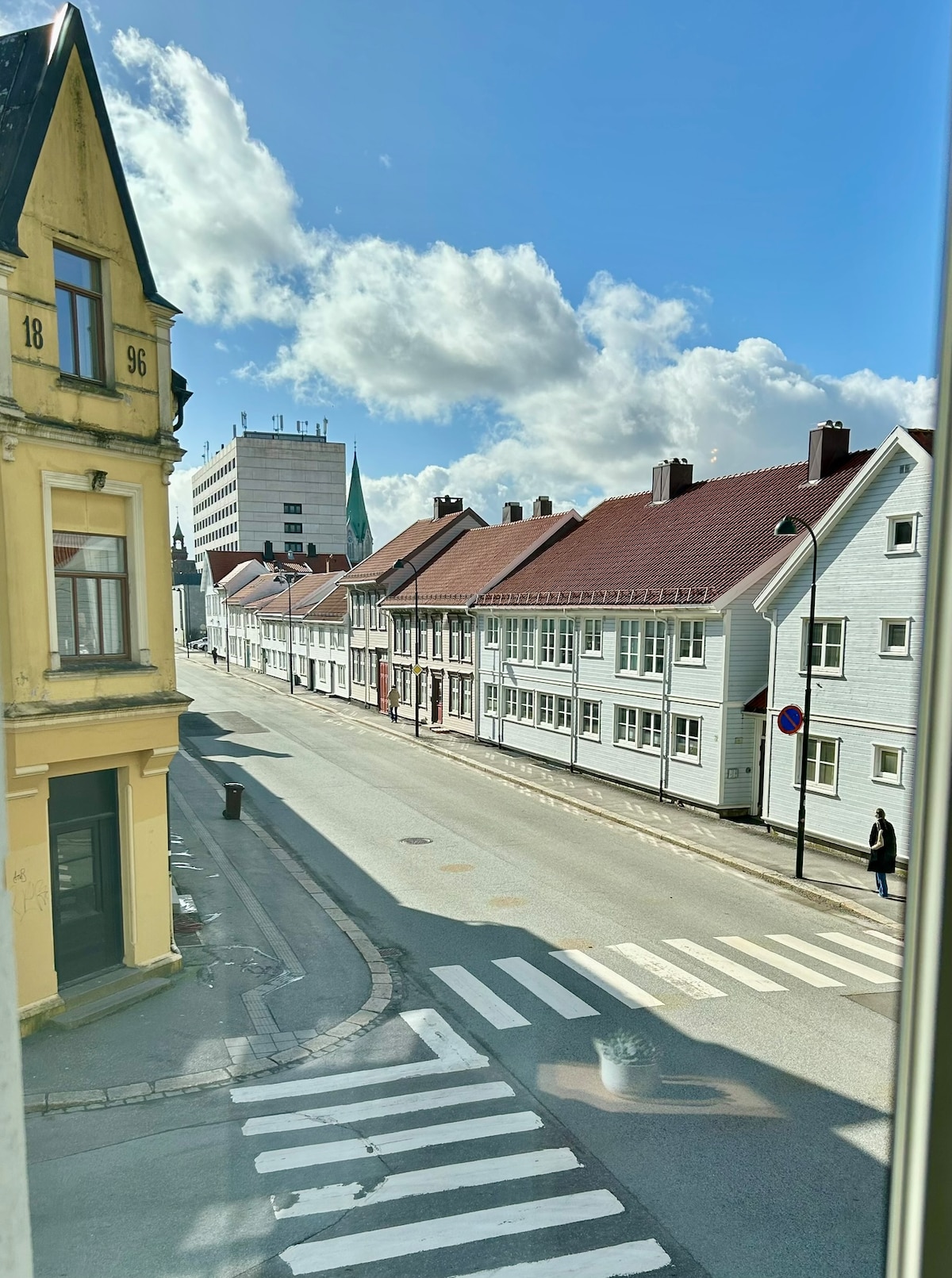 A view looking down a quiet street reveals a mix of traditional wooden buildings and modern architecture under a partly cloudy sky. A pedestrian is visible in the distance, and a zebra crossing leads across the road, enhancing accessibility.