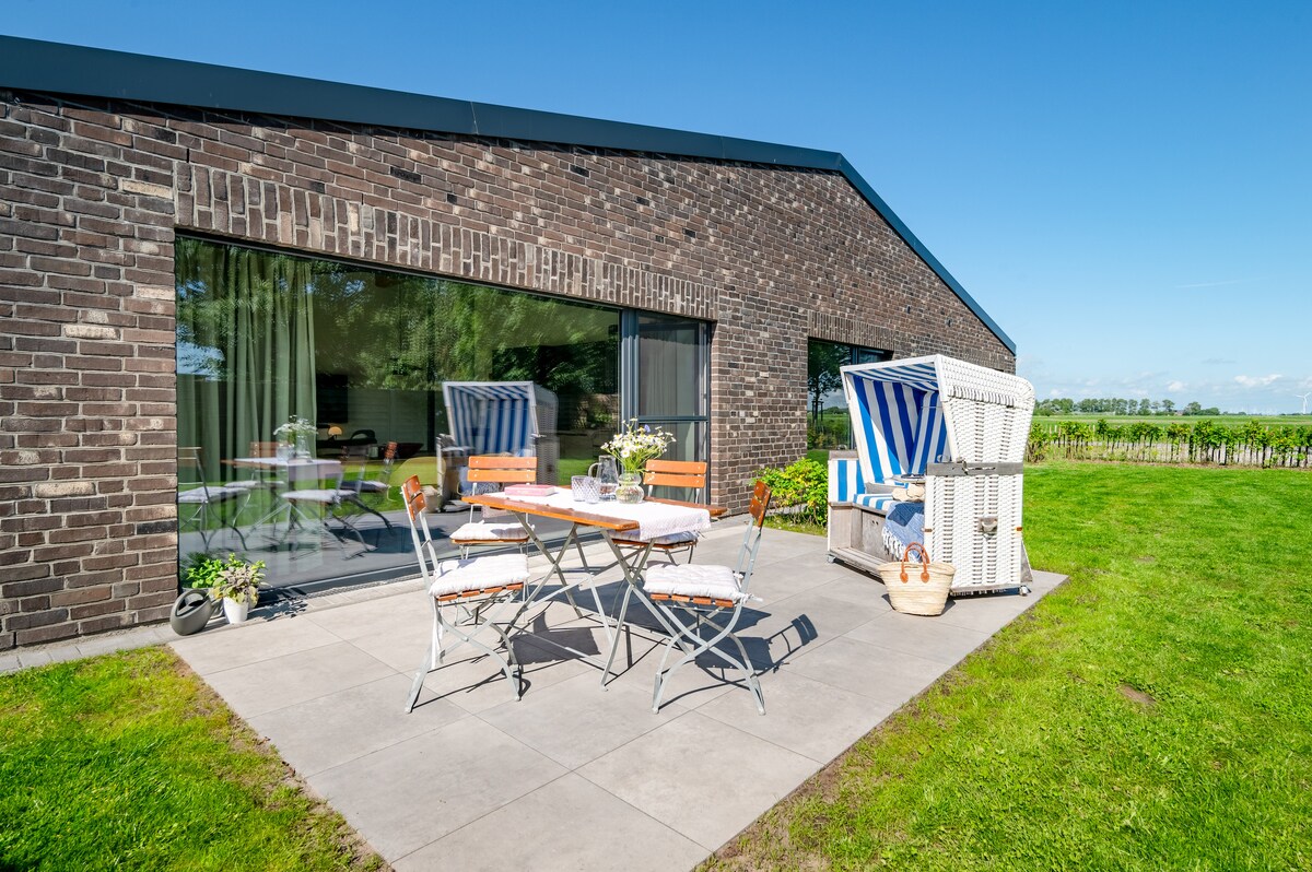 An outdoor patio area features a dining table set for four, accompanied by comfortable folding chairs. A beach chair with blue and white stripes adds a relaxed touch. The backdrop includes a modern brick facade and a well-maintained lawn, with clear skies enhancing the peaceful setting.