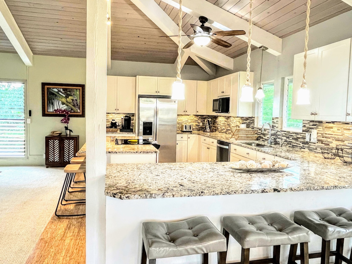 A spacious kitchen is presented with modern amenities and granite countertops. Light fixtures hang above the island bar with seating for four, while stainless steel appliances and a decorative tile backsplash enhance the design. Natural light filters through large windows, creating a bright environment.