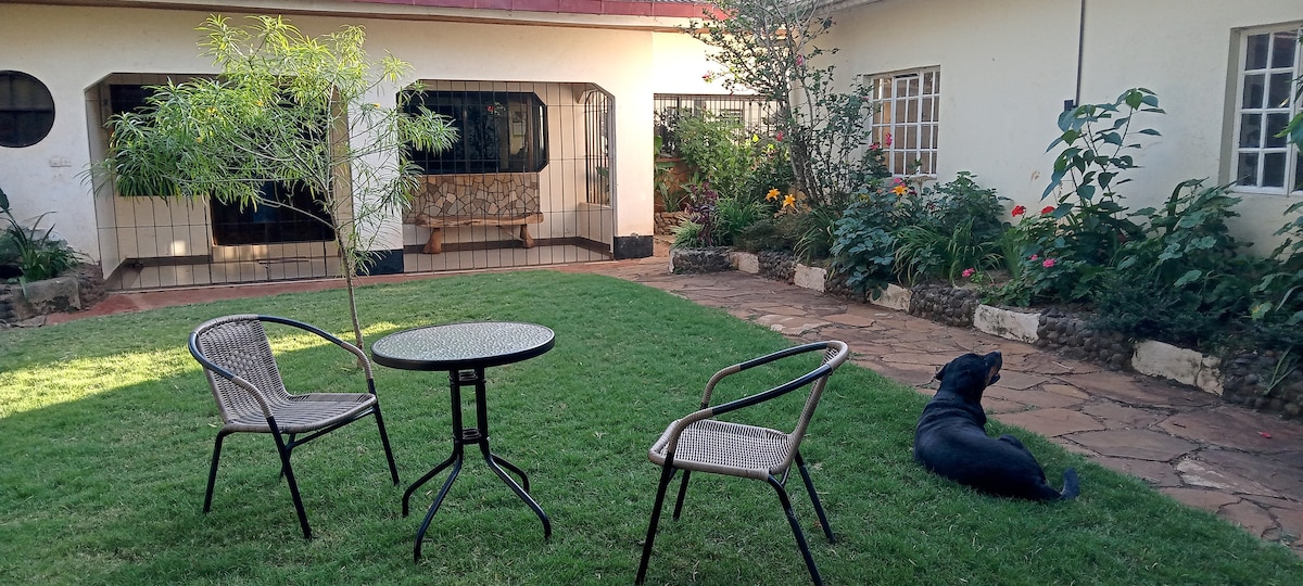 A private patio area features a small table surrounded by two chairs on a grassy lawn. A black dog rests nearby, with vibrant flowers and lush greenery framing the scene. The patio provides a view of the guesthouse's inviting living space through large, arched windows.