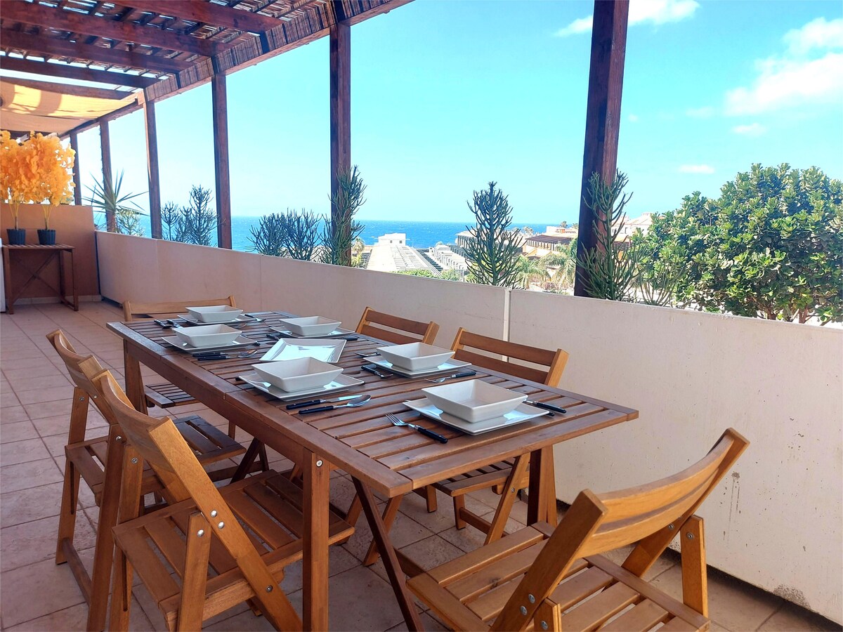 A spacious terrace is featured, furnished with a wooden dining table set for eight. Ocean views are visible in the background, along with greenery from nearby plants. Sunlight illuminates the area, enhancing the outdoor dining experience.