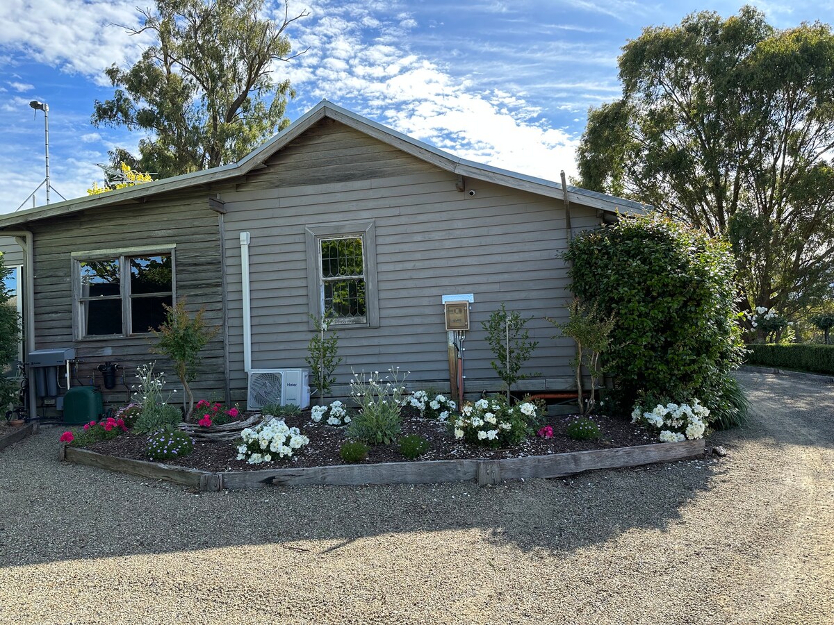 The exterior of the cabin is shown, featuring a rustic wooden façade, framed by a landscaped area with flower beds that include white and pink blooms. A gravel pathway leads to the entrance, and windows provide glimpses of natural light inside.