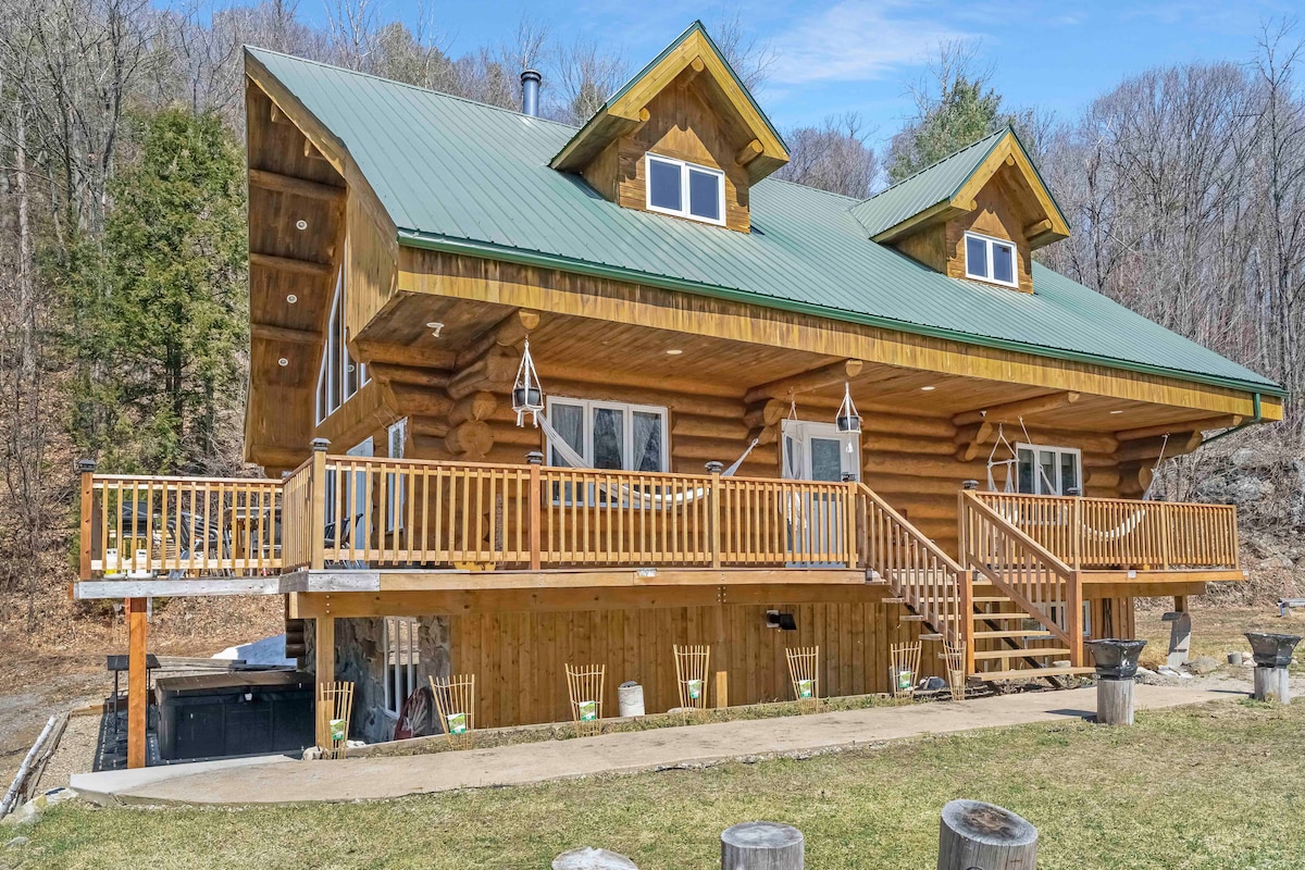 The exterior of the log chalet showcases a spacious deck with a green metal roof and large wooden accents. Symmetrical gables and multiple front-facing windows allow for abundant natural light. Stairs lead to both the upper and lower deck areas, providing easy outdoor access.
