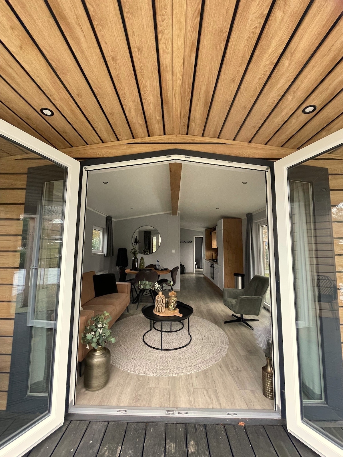 A welcoming entrance is seen through double doors, revealing a bright open living space furnished with a comfortable sofa and armchair. A round coffee table sits in the center, surrounded by natural light from large windows and highlighting warm wooden accents throughout.