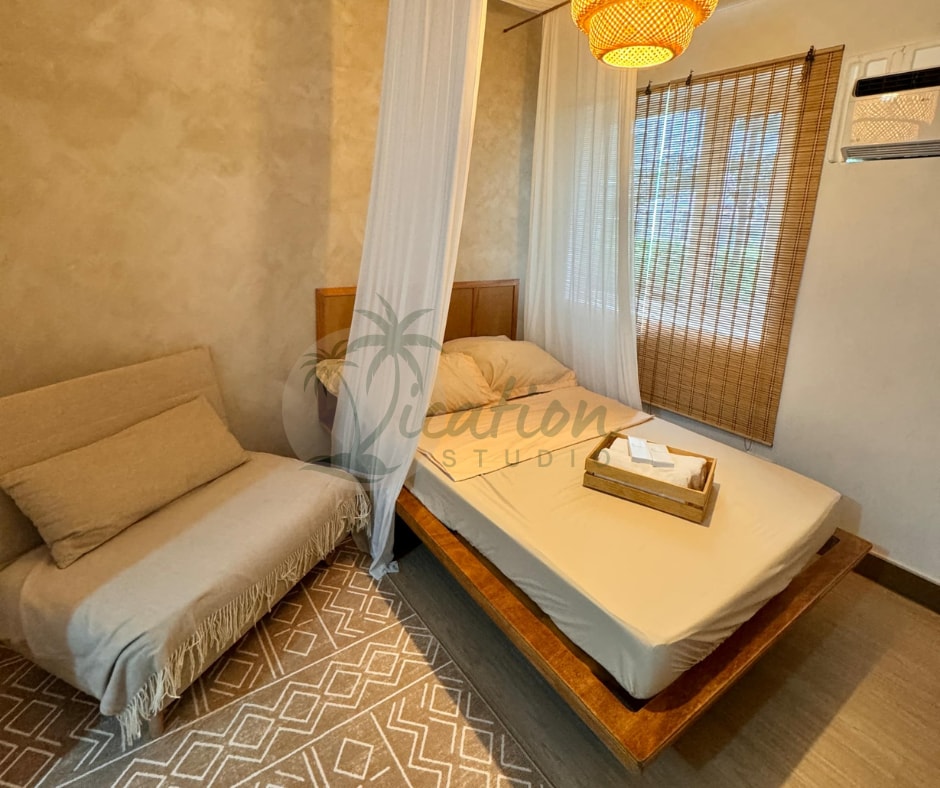 A serene sleeping area is highlighted by a spacious bed draped with a soft canopy. A cozy sofabed is positioned nearby, complemented by a woven area rug. Natural light filters through sheer curtains, creating a calm and inviting ambiance.