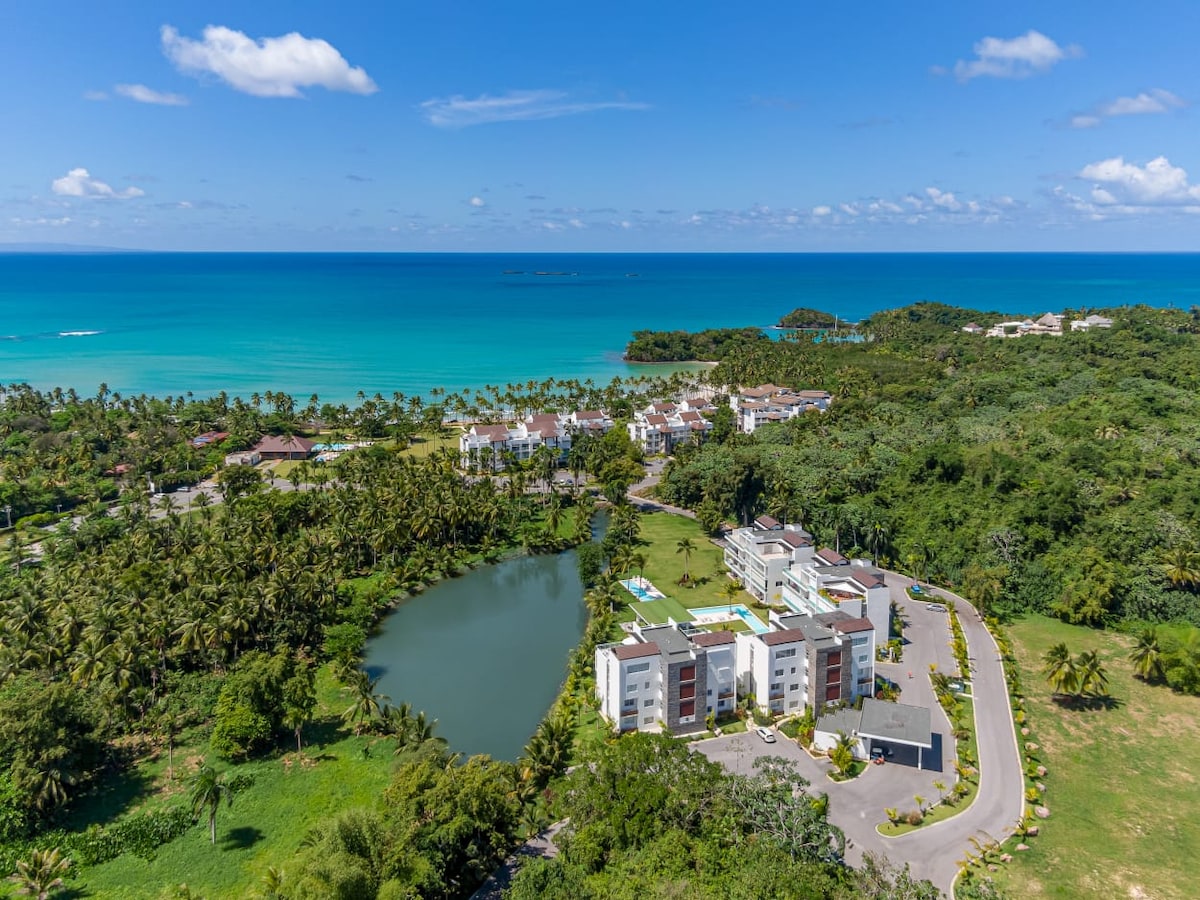 An aerial view of the Playa Bonita Beach Residences showcases their proximity to both the serene ocean and lush greenery. The image highlights a calm lagoon surrounded by palm trees, with the vibrant blue waters of the sea visible in the background.