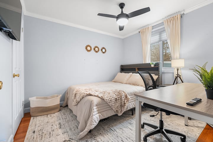 Dormitorio luminoso y acogedor con una cómoda cama tamaño queen, ventilador de techo, escritorio y televisor montado en la pared, perfecto para relajarse o trabajar cómodamente.