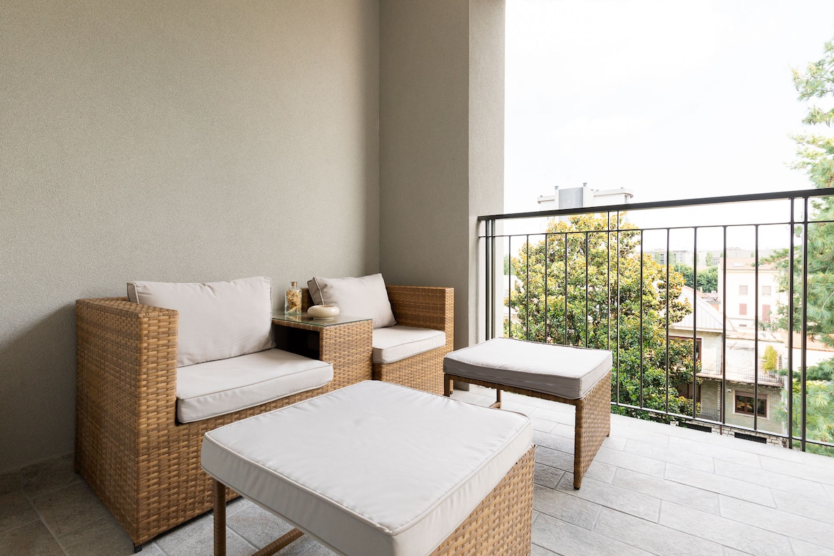 An outdoor balcony space features a comfortable seating area with two wicker chairs and a low table, all adorned with soft cushions. The balcony overlooks greenery, offering a peaceful environment for relaxation.