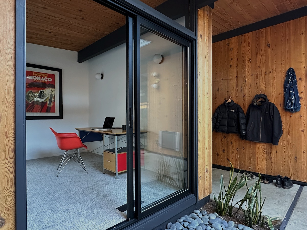 An office space is visible through glass doors, featuring a modern desk with a computer and a red chair. The exterior showcases wooden paneling and a stone pathway, complemented by hanging jackets and a framed art piece on the wall.
