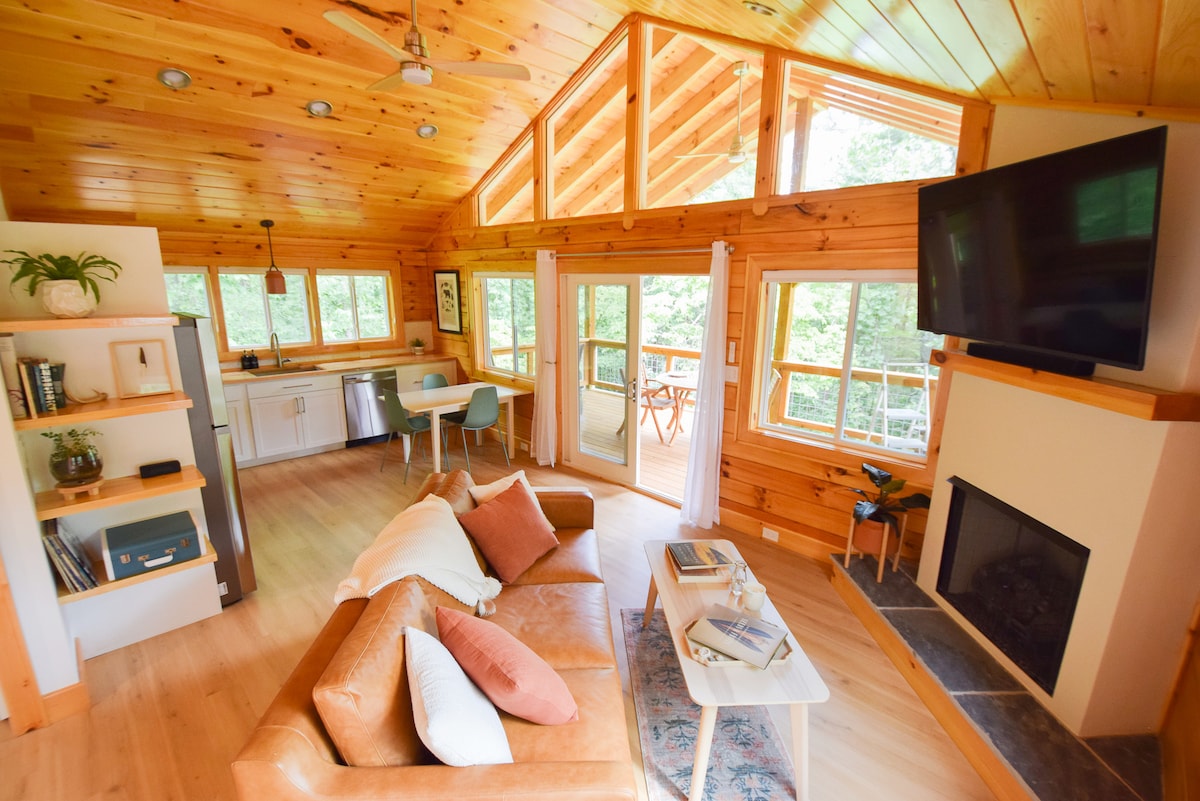 The living area features a vaulted ceiling and large windows that invite natural light. A cozy leather sleeper sofa is arranged around a coffee table, with a fireplace and a wall-mounted television nearby. The open-plan space extends to the kitchen and dining area.