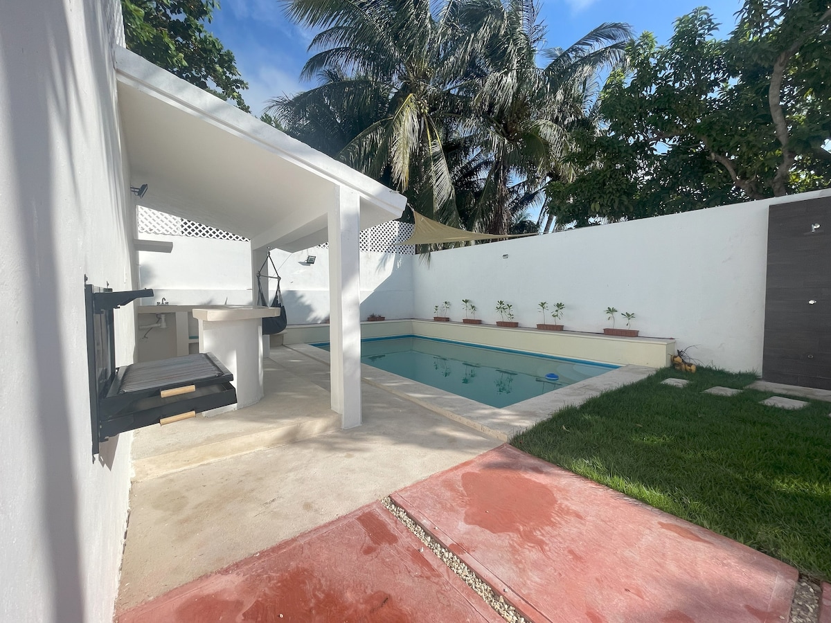 An outdoor area is showcased, featuring a swimming pool surrounded by a neatly maintained garden. A shaded bar area is presented with seating and a grill. Lush palm trees are visible in the background, enhancing the space's tropical feel.