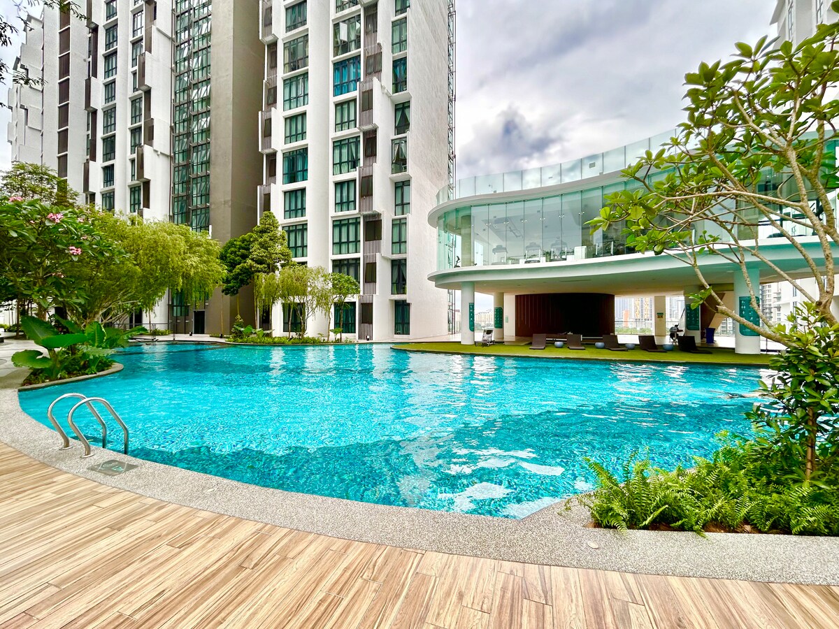 A large swimming pool is set within a landscaped area surrounded by modern residential buildings. Lush greenery and seating areas are visible around the pool, creating a serene environment. The water glistens under overcast skies, inviting relaxation.