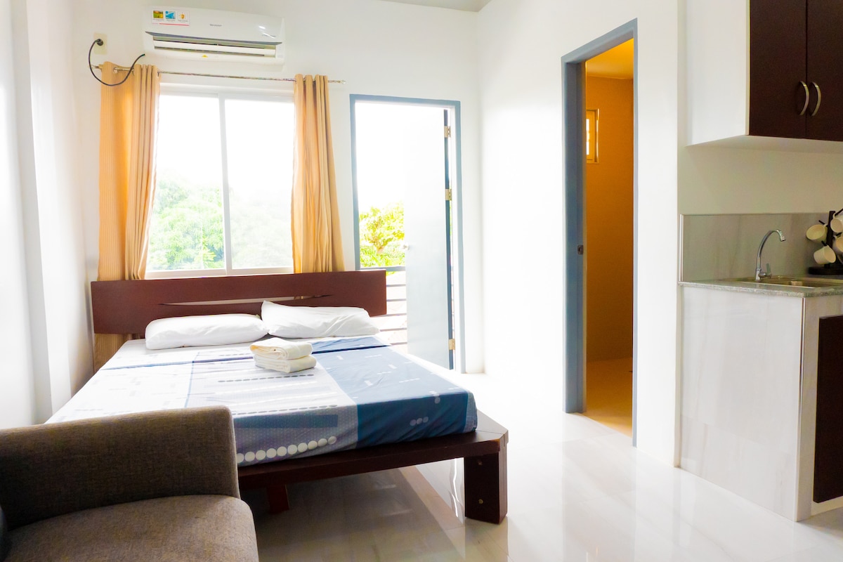 A well-lit bedroom features a spacious bed with light linens and a cozy sofa positioned nearby. Large windows provide natural light and views of greenery outside. A small kitchenette is visible in the corner, and a door leads to another room, enhancing the space's functionality.