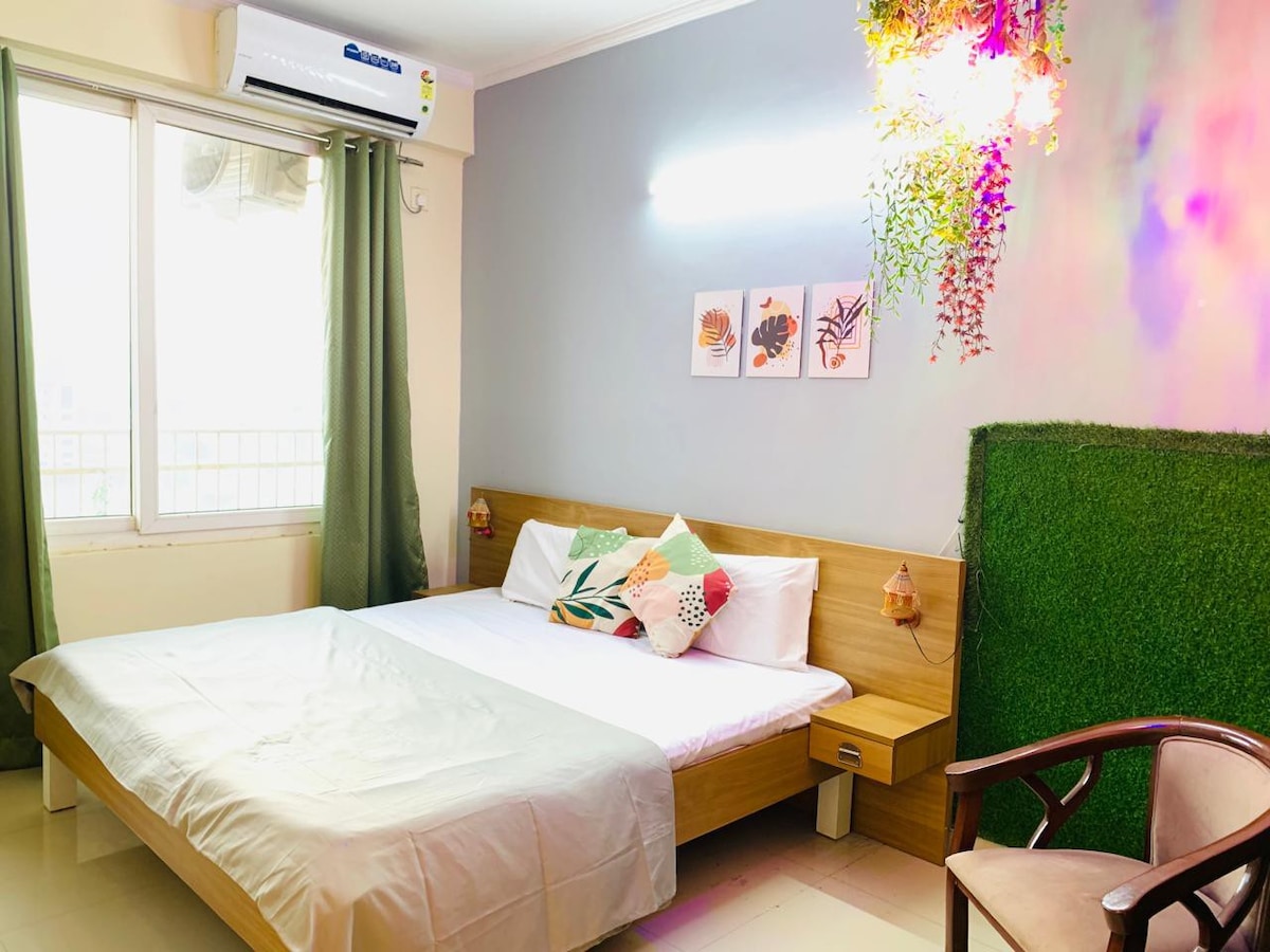 A spacious sleeping area is highlighted by a king-size bed with a comfortable mattress and decorative pillows. Natural light flows through the large window, adorned with green curtains. Wall art featuring plant motifs adds a touch of style, complemented by vibrant hanging decor.