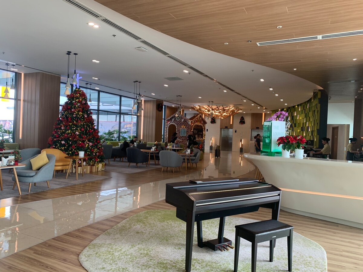 A spacious lounge area is visible with a grand piano at the foreground. Holiday decor includes a decorated Christmas tree. Comfortable seating arrangements are positioned throughout, featuring a mix of armchairs and sofas. Natural light enters through large windows, enhancing the inviting atmosphere.