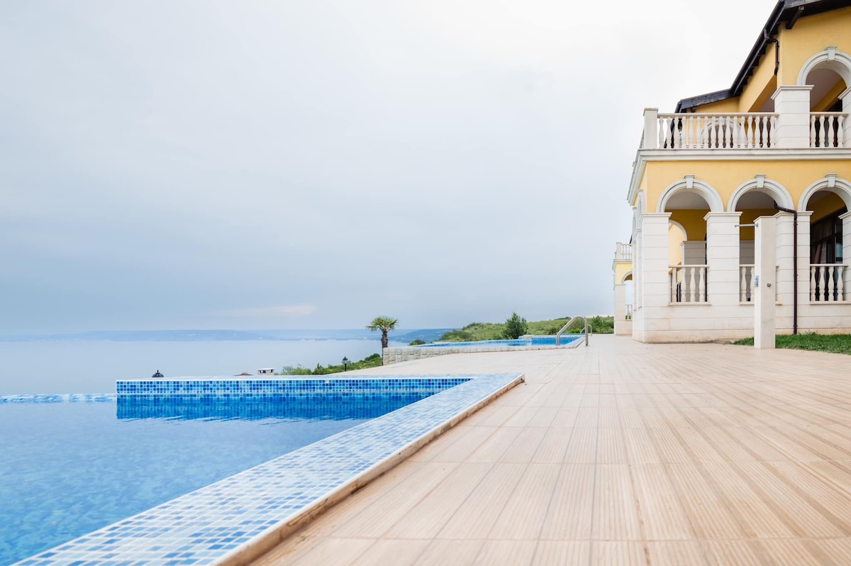 The villa's exterior showcases a spacious terrace made of natural wood, leading to a heated infinity pool that blends seamlessly with the sea view. The well-designed architecture features arched columns and large doors, complementing the serene landscape.
