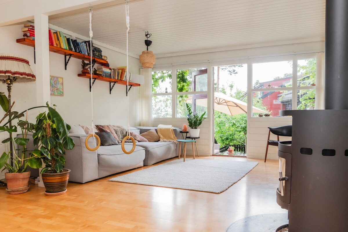 A bright and spacious living area is complemented by large windows allowing natural light to illuminate the space. A cozy couch is surrounded by plants, with decorative shelves filled with books above. A simple rug covers the wooden floor, while a wood-burning stove adds warmth.