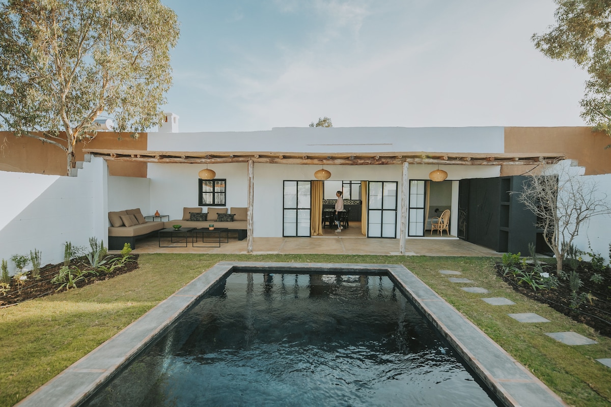 An outdoor view of the villa highlights a spacious patio area with comfortable seating. A tranquil pool reflects the sky, surrounded by well-maintained greenery. The wooden beams and pendant lights above add an inviting touch to the open space, accessible from the living area.