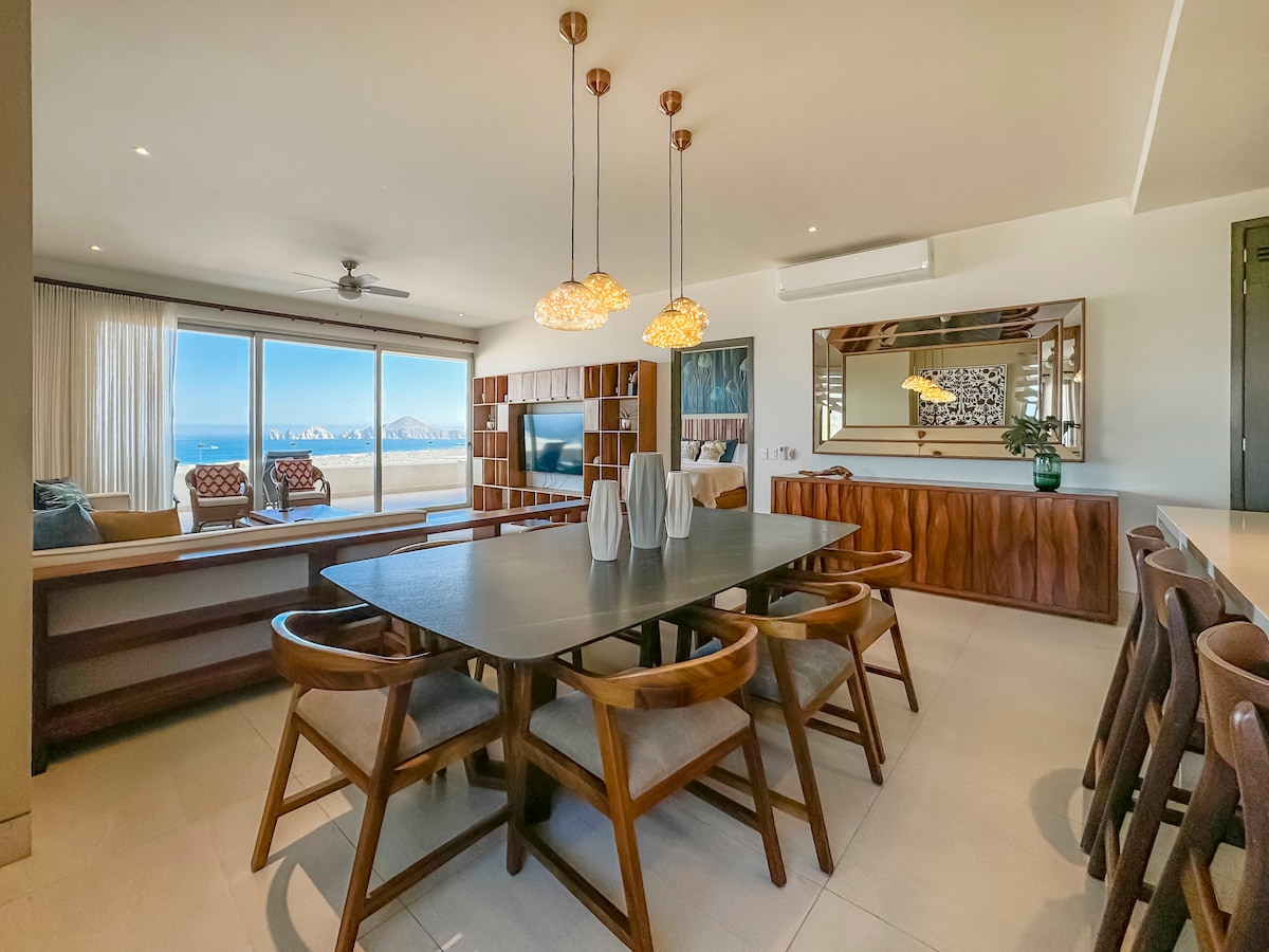 A modern dining area features a large table surrounded by wooden chairs. Natural light floods the space through expansive windows, offering views of the ocean. A comfortable seating area with two patterned chairs is visible in the background, alongside a stylish entertainment unit.