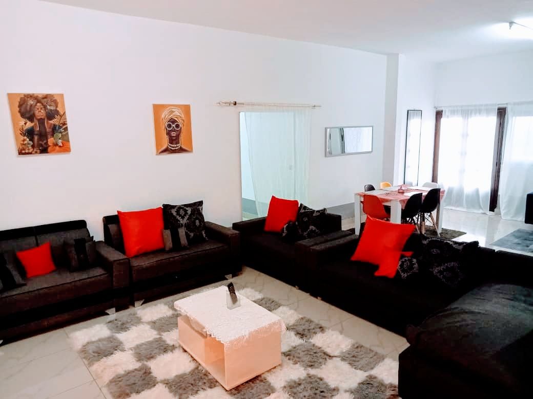 A spacious living area features several dark sofas adorned with red accent pillows, arranged around a light-colored coffee table. A soft area rug with a neutral tone lies beneath. Natural light brightens the space, with dining chairs and a table visible in the background.