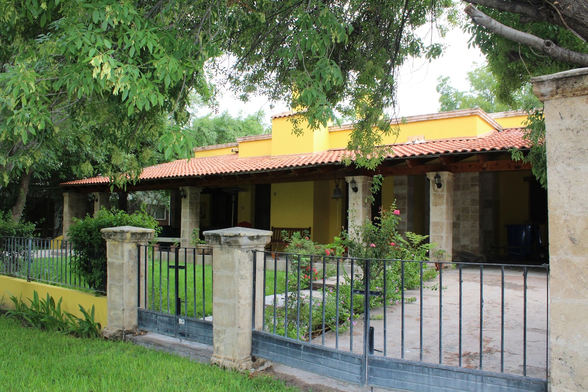 A charming yellow house with a red-tiled roof is surrounded by lush greenery. The front porch features stone columns and a spacious entryway, while a manicured lawn frames the property, enhancing its inviting presence.
