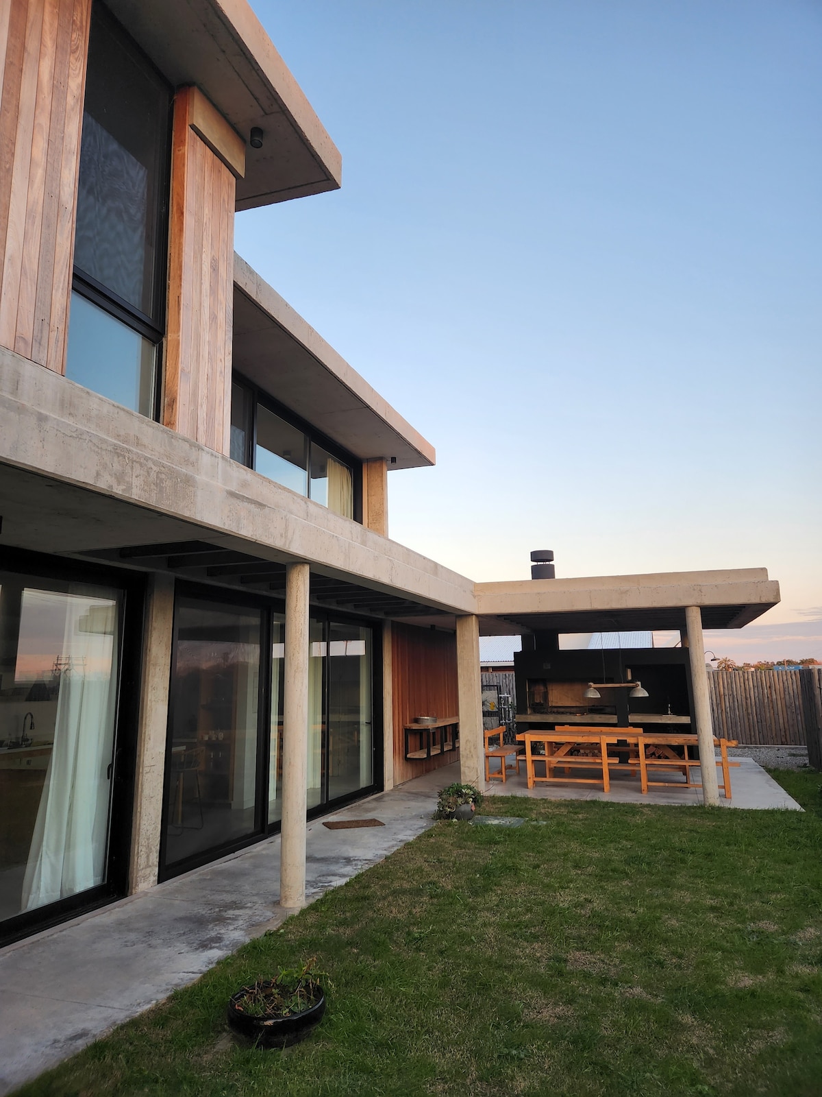 The exterior of a modern house is presented, featuring a mix of concrete and wooden elements. A spacious outdoor dining area with a large table and a grill is visible. The lawn is well-maintained, with the structure's large glass doors reflecting the evening light.