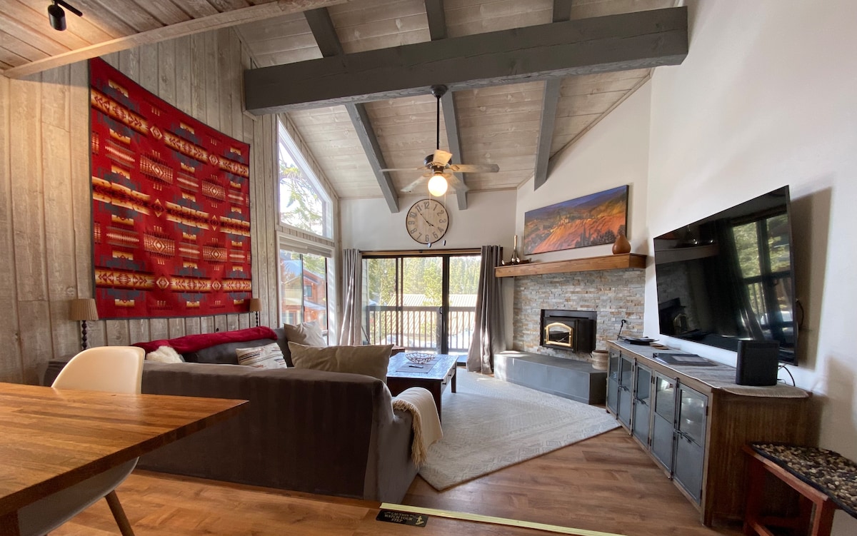 A spacious living area features a large sectional sofa and a wooden coffee table. Natural light floods in through the large windows, highlighting decorative elements like a vibrant wall hanging and a clock. A stone fireplace adds a comfortable focal point to the room.