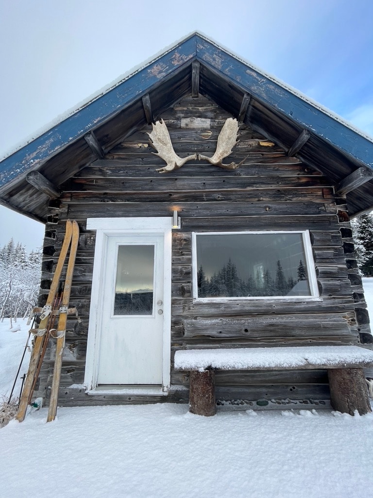 The front exterior of a rustic log cabin is presented, featuring a simple door and a large window. A bench made of logs is positioned beneath the window. Mounted antlers adorn the overhanging roof, while snow covers the ground and surrounding landscape.