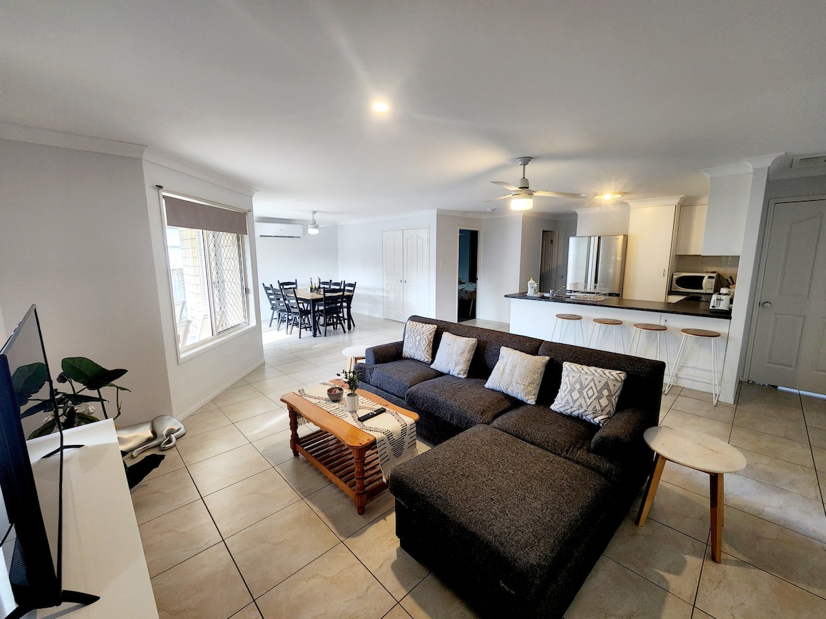 A spacious living area showcases a dark sectional sofa with a coffee table in front. Natural light enters through the large window, and a dining area with a wooden table and chairs is visible in the background. The kitchen features contemporary cabinetry and appliances.