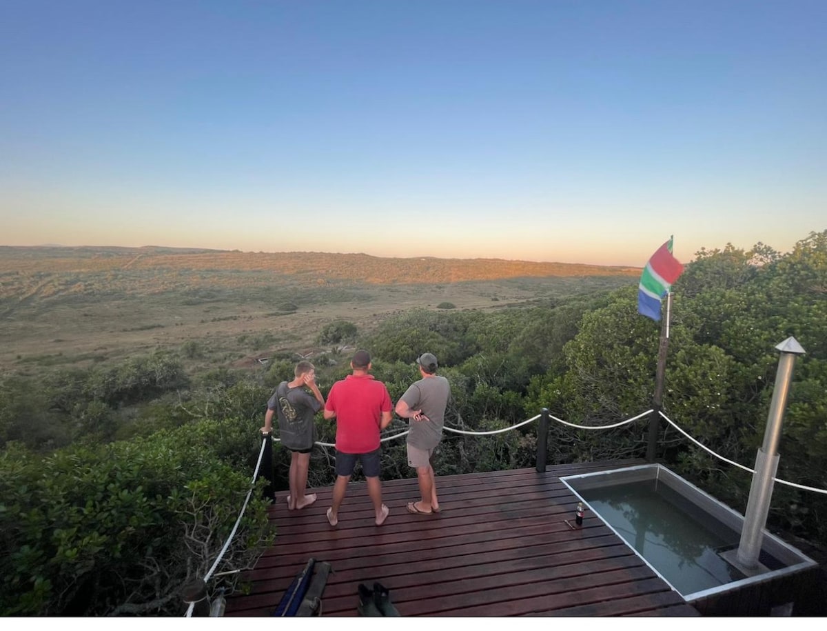 A private deck overlooks a vast landscape at sunset, with three individuals standing together. A small hot tub is positioned to one side, and a flag is displayed nearby, indicating inclusivity. Lush greenery surrounds the area, enhancing the serene atmosphere.