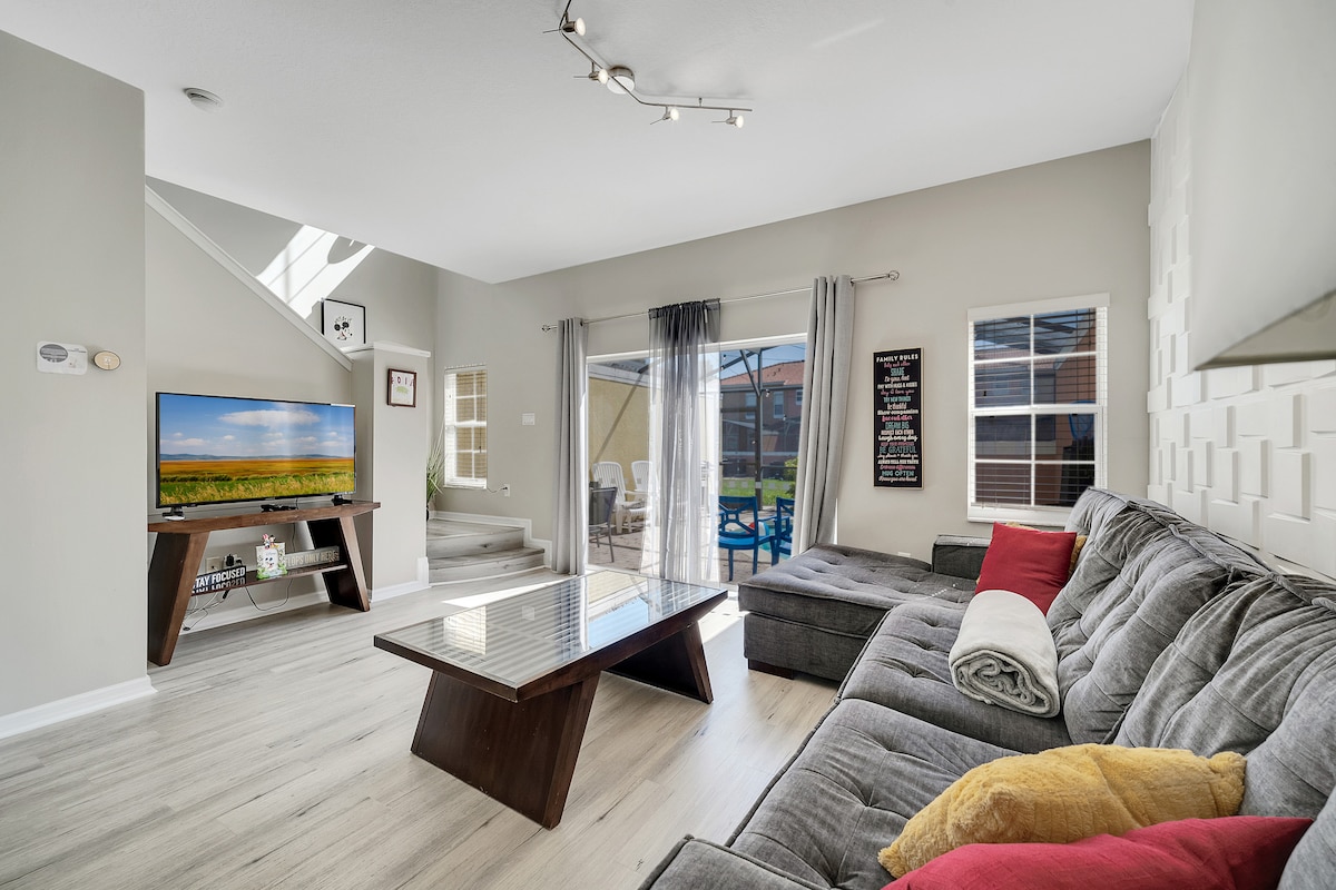 The spacious living room showcases a large, gray sectional sofa positioned around a sleek coffee table. A 50-inch smart TV is visible on a media unit. Natural light filters through the sliding glass doors, providing views of the screened patio and private pool area.