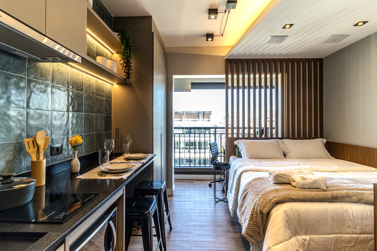 A modern studio space features a well-equipped kitchenette with sleek appliances and bar stools. A spacious bed is positioned against a textured wall, while natural light enters through a balcony door. The decor combines functionality with stylish design elements.