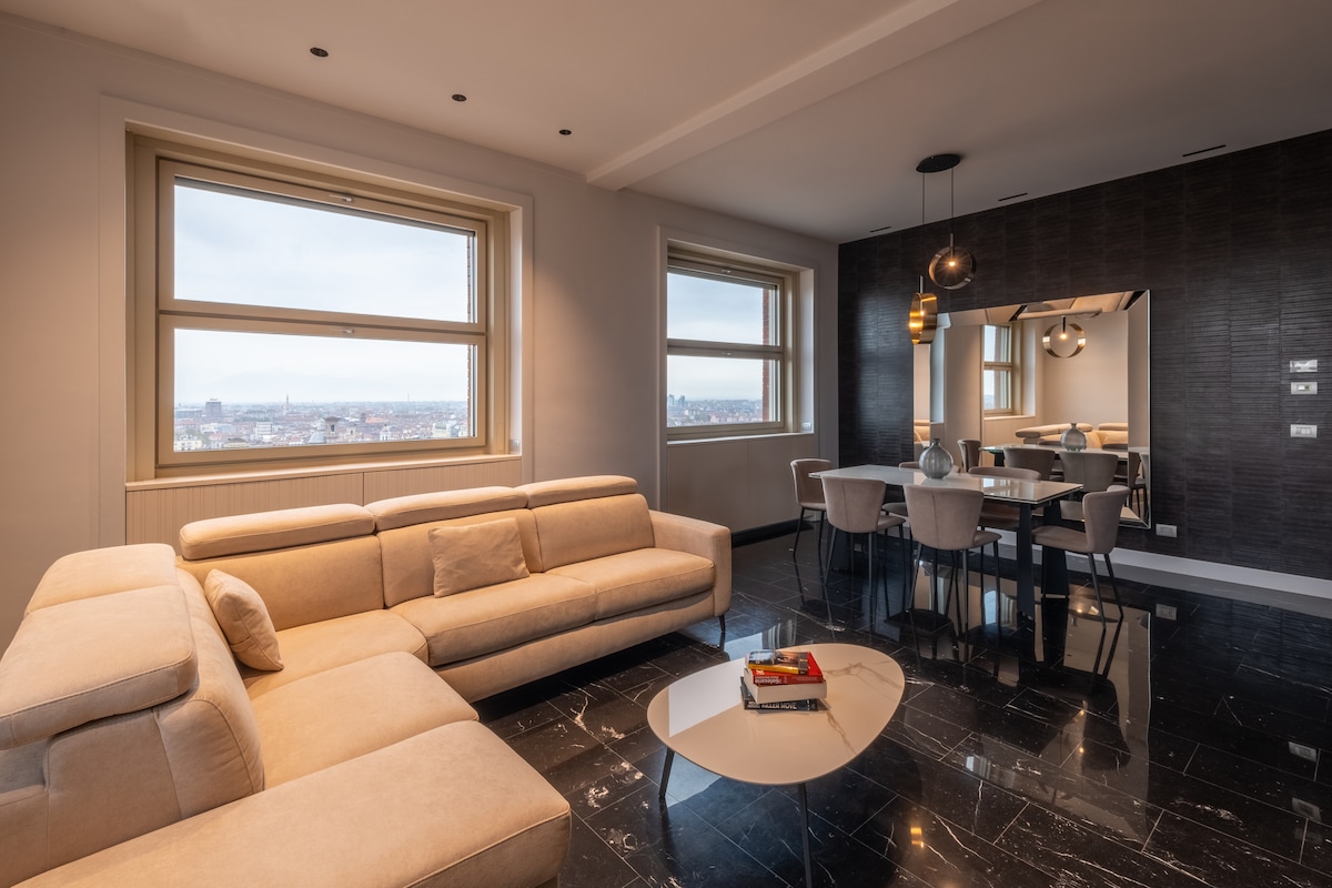 A modern living area is displayed, featuring a spacious light-colored sectional sofa and a sleek coffee table. Large windows offer expansive city views. A dining table with chairs and decorative lighting enhance the contemporary design, while dark flooring adds a touch of elegance.