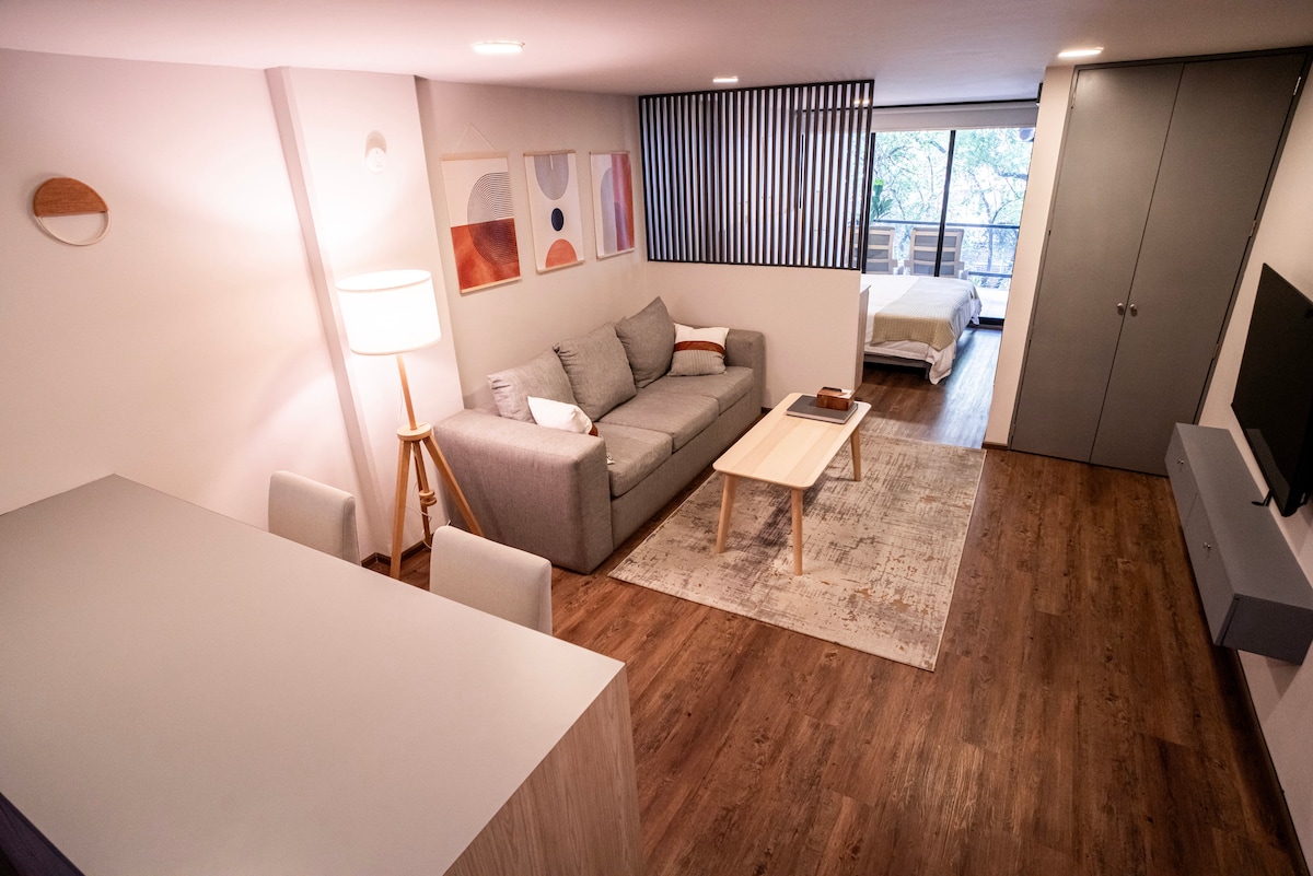 An open living area features a light gray sofa paired with a natural wood coffee table on a neutral area rug. A dining space with two chairs is visible, along with art pieces hanging on the wall. Floor-to-ceiling windows allow natural light, revealing a glimpse of the bedroom.