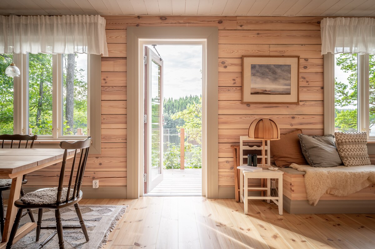 Bright interior space features natural wood paneling and large windows opening to a scenic view. A dining table is paired with black chairs, while a cozy seating area is accented with cushions and a soft throw. The doorway leads to an outdoor deck surrounded by greenery.