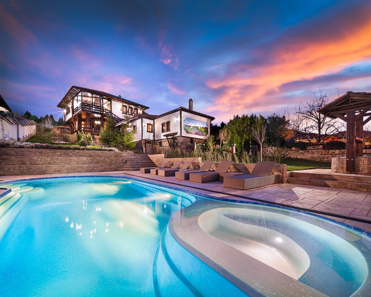 A private heated pool is illuminated with soft blue light, surrounded by lounge chairs nestled on the stone patio. A twilit sky features streaks of pink and orange, complementing the rustic architecture of the villa in the background, framed by landscaped greenery.