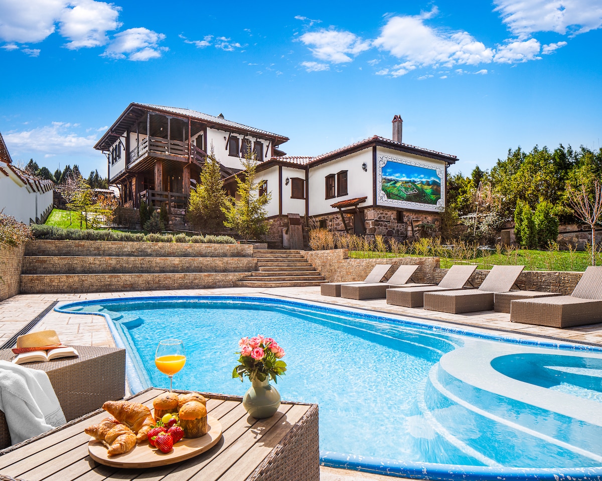 A private heated pool is featured in the foreground, accompanied by a wooden dining table set with a breakfast spread. The villa stands in the background, showcasing a blend of traditional architecture and modern elements, with expansive outdoor spaces bathed in natural light.