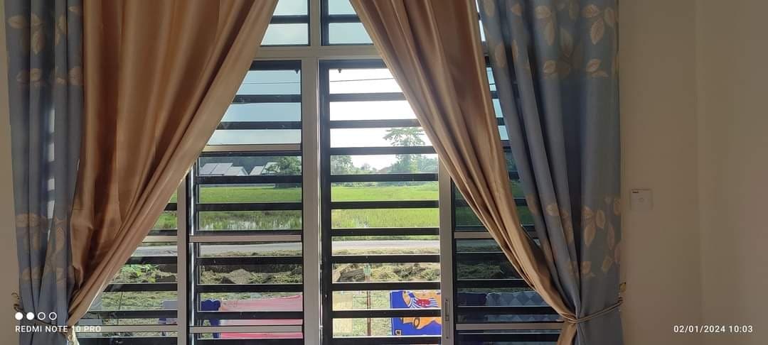 A view from a large window, framed by light blue and beige curtains. Lush green rice fields are visible outside, creating a serene backdrop. The window features horizontal slats, allowing natural light to filter in while maintaining privacy.
