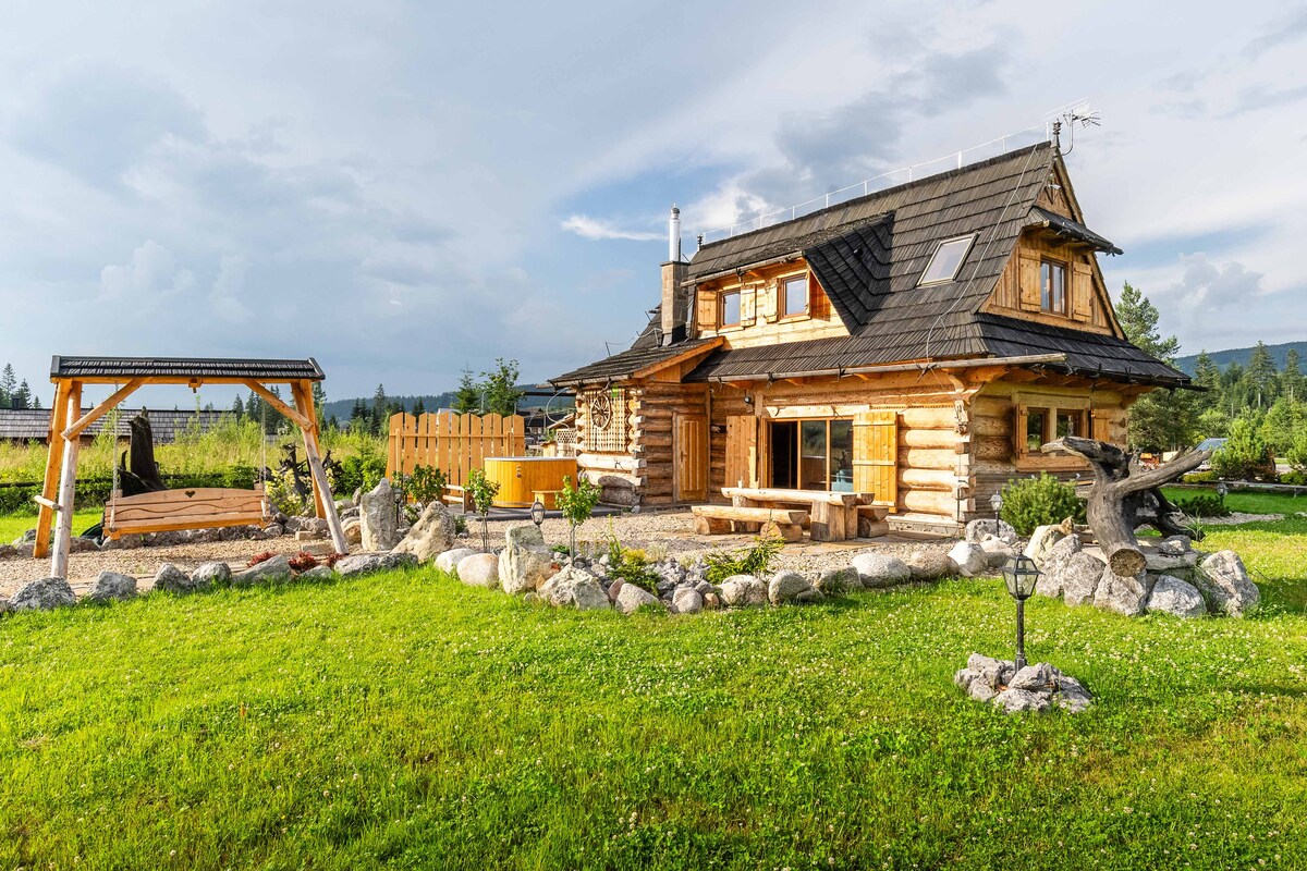 A charming log cabin is set against a backdrop of greenery, featuring a wooden swing set nearby. A wraparound porch includes a spacious seating area, while a stone path leads through a landscaped garden. Natural elements and rustic architecture create a serene outdoors.