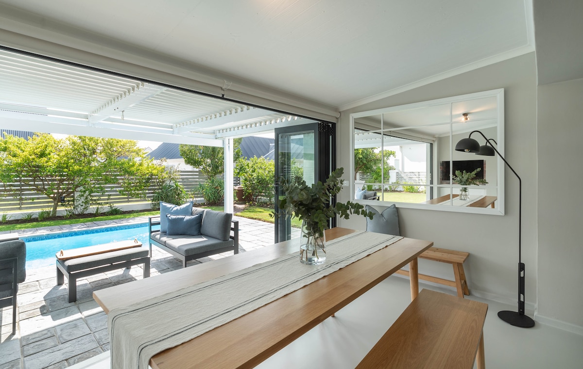 A modern dining area is highlighted by a long wooden table, seating several guests, with a light-colored runner adding a subtle touch. Large glass doors provide a view of the private pool and garden, enhancing the indoor-outdoor connection.