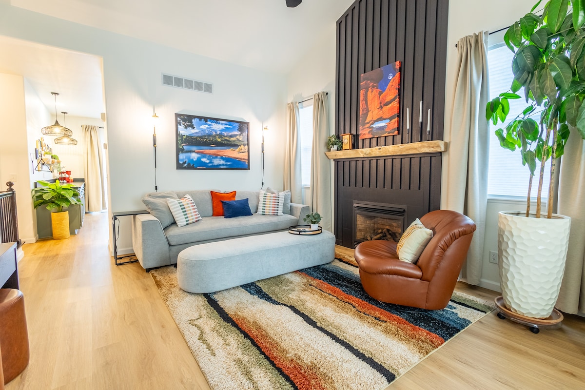 A modern living room features a large, L-shaped gray sofa adorned with patterned throw pillows. A unique circular coffee table sits on a colorful area rug, while a brown leather armchair is positioned nearby. A wall-mounted artwork and a fireplace are focal points, complemented by soft lighting and natural plant decor.
