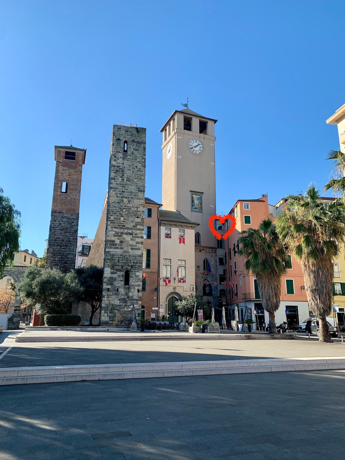 An historic square is highlighted by a clock tower and accompanying stone structures. Palm trees frame the foreground, while colorful buildings line the square, adding charm to the setting. Clear blue skies create a bright backdrop, emphasizing the inviting atmosphere of the area.