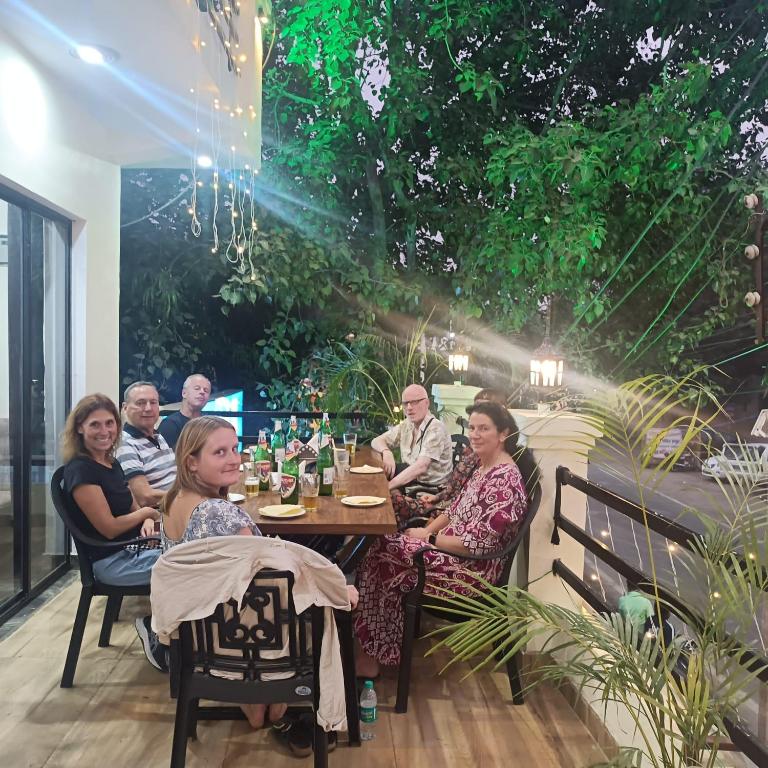 A communal dining area features a long table set with plates and drinks. Guests are gathered, engaged in conversation. Lush greenery surrounding the space adds a refreshing touch, while soft lighting creates a relaxed evening atmosphere.