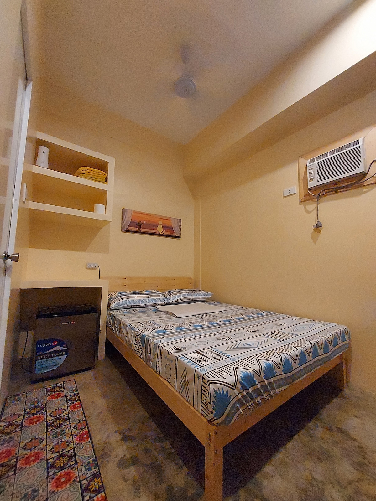 A cozy studio apartment is depicted, featuring a full double bed with patterned bedding. A compact refrigerator is positioned near the bed. Wall-mounted storage holds towels and essentials. Soft yellow walls and a ceiling fan contribute to the comfortable ambiance.