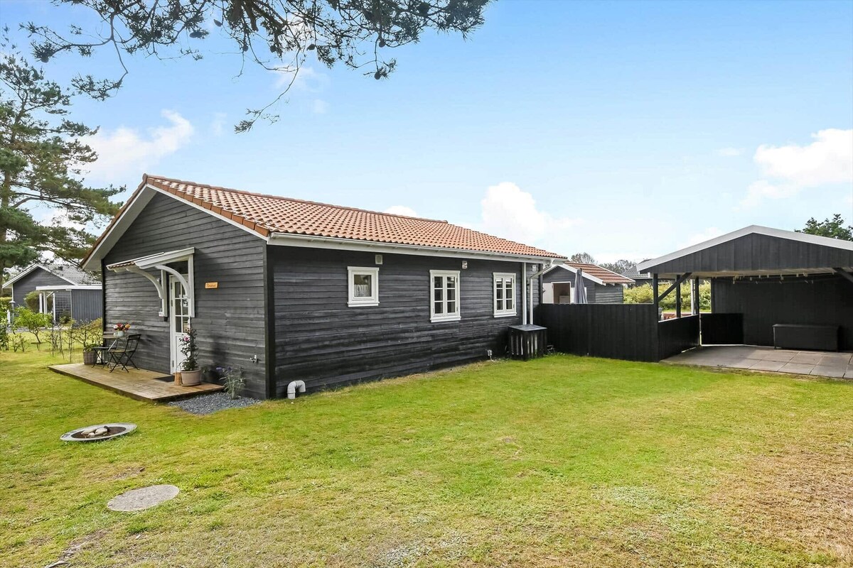 The exterior of a charming black wooden house is depicted, featuring a well-maintained lawn. A carport is present beside the house, providing covered parking. Several windows allow natural light to enter, while a small seating area with a table and chairs is visible on the porch.